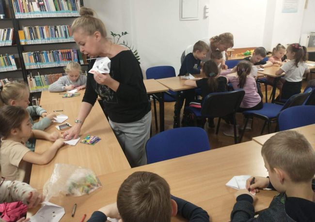 Biblioteka. Dzieci siedzą przy stolikach wykonując kotki i pieski metodą origami zgodnie z poleceniami prowadzącej.