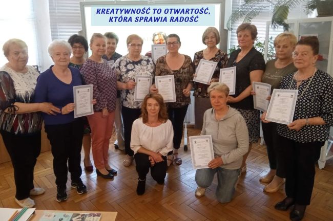 Zdjęcie grupowe. Seniorzy stoją, trzymają certyfikaty ukończenia warsztatów.