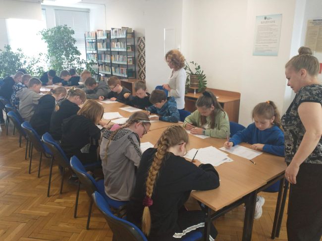 Uczniowie siedząc przy stolikach wykonyją zadania. Obok uczniów nauczyciele bibliotekarze
