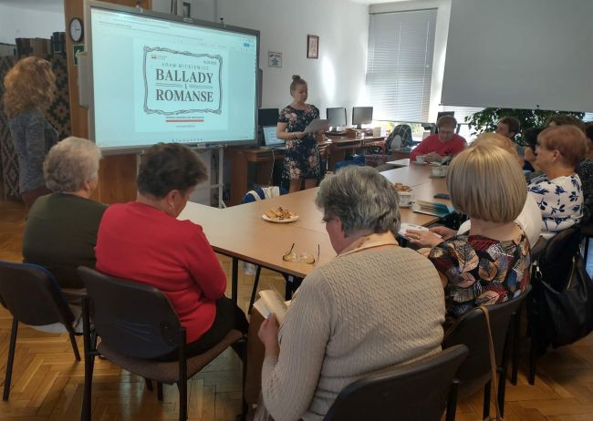 Prowadzące przedstawiają „Ballady i romanse” Adama Mickiewicza . Nawiązują do  tematu Narodowego Czytania w 2022 roku. Słuchacze Uniwersytetu Trzeciego Wieku w Tomaszowie Lubelskim siedzą przy stołach.