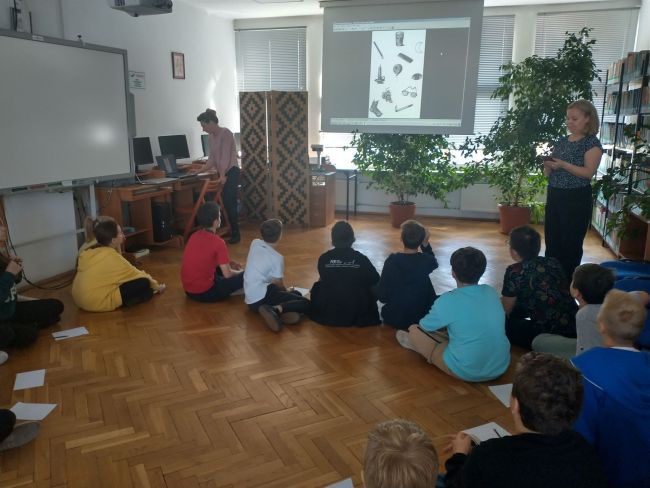 Na zdjęciu uczniowie siedzący w kręgu. Obok prowadzący zajęcia nauczyciele bibliotekarze. W tle na ekranie projektora wyświetlane są zadania dla uczniów 