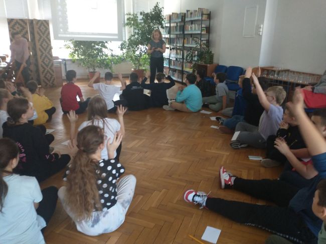 Na zdjęciu uczniowie siedzący w kręgu. Obok prowadzący zajęcia nauczyciele bibliotekarze. W tle na ekranie projektora wyświetlane są zadania dla uczniów 
