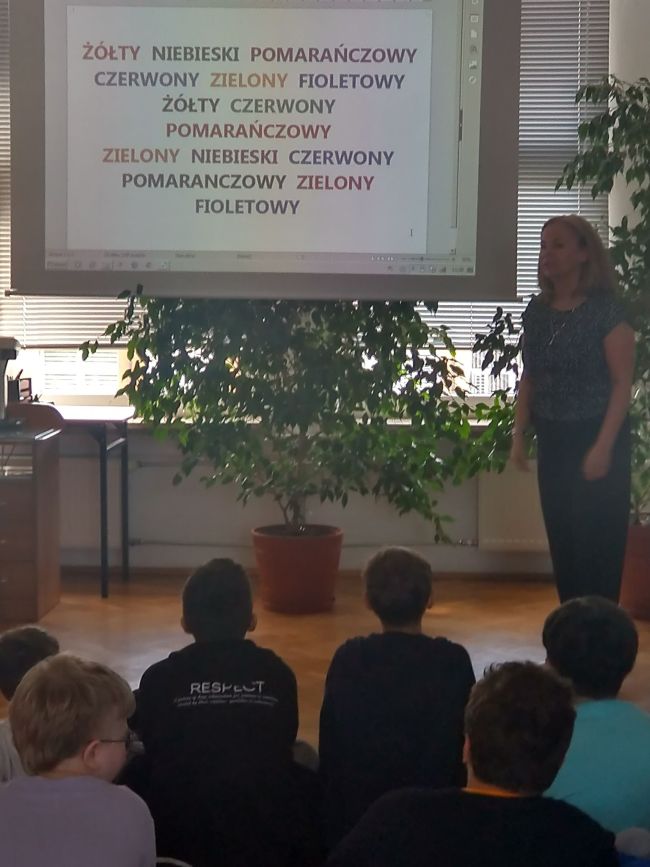 Na zdjęciu uczniowie siedzący na podłodze. Obok prowadząca zajęcia. Na ekranie projektora wyświetlane są zadania dla uczniów 