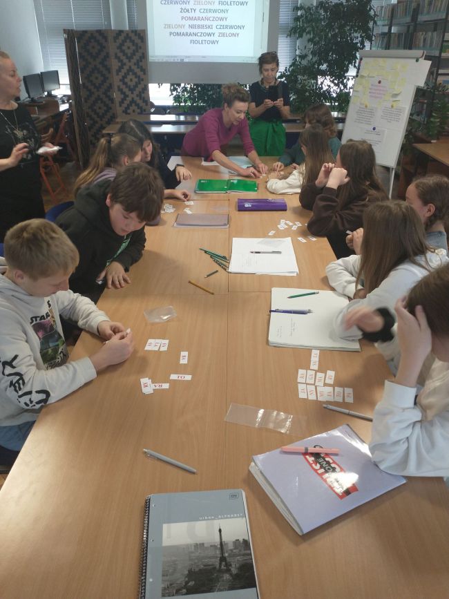 Na zdjęciu uczniowie siedzący przy stolikach oraz prowadzący zajęcia nauczyciele bibliotekarze.  Uczniowie wykonują zadania przygotowane przez nauczycieli. W tle na ekranie  projektora wyświetlane są ćwiczenia dla uczniów, obok flipchart z tytułem zajęć