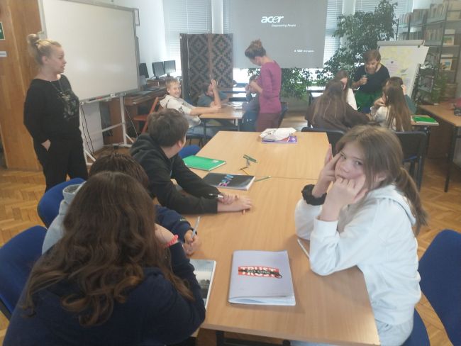 Na zdjęciu uczniowie siedzący przy stolikach oraz prowadzący zajęcia nauczyciele bibliotekarze. Uczniowie wykonują zadania przygotowane przez nauczycieli.  
