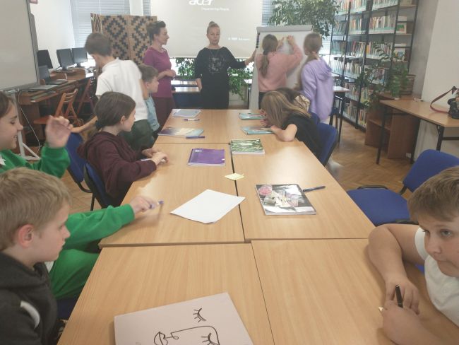  Na zdjęciu uczniowie siedzący przy stolikach oraz prowadzący zajęcia nauczyciele bibliotekarze. Część uczniów przykleja do flipcharta karteczki z odpowiedziami na pytanie zadane przez nauczycieli