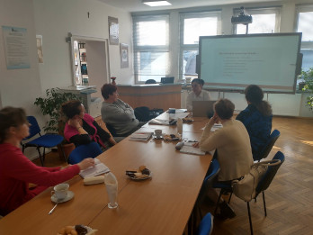 Czytelnia biblioteki pedagogicznej. Uczestnicy warsztatów siedzą przy stołach. W tle tablica interaktywna, na której wyświetlana jest prezentacja.