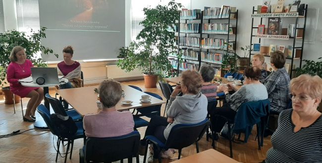 Słuchacze Uniwersytetu Trzeciego Wieku siedzą przy stolikach. Prowadzące udostępniają prezentację o Wisławie Szymborskiej