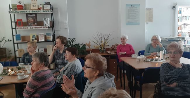 Słuchacze Uniwersytetu Trzeciego Wieku siedzą przy stolikach. Prowadzące udostępniają prezentację o Wisławie Szymborskiej