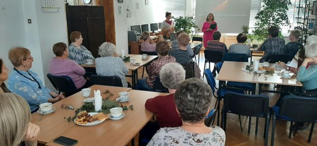 Słuchacze Uniwersytetu Trzeciego Wieku siedzą przy stolikach. Prowadzące udostępniają prezentację o Wisławie Szymborskiej