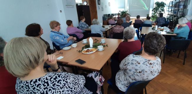 Słuchacze Uniwersytetu Trzeciego Wieku siedza przy stolikach. Prowadzące udostępniają prezentację o Wisławie Szymborskiej