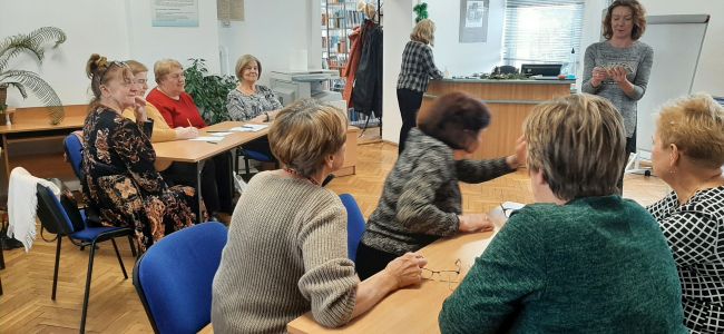Seniorzy grają w Kalambury. Prowadząca pokazuje karty z hasłem do wyboru.