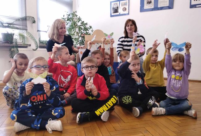 Zdjęcie grupowe z przedszkolakami. Siedzą na podłodze i prezentują pokolorowane przez siebie motyle.