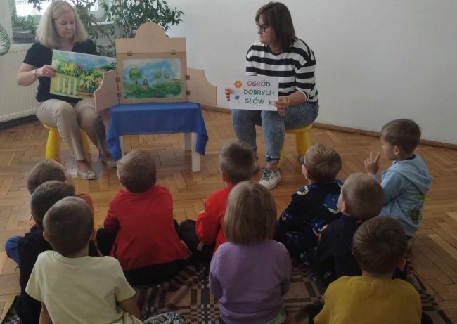 Prowadzaca prezentuje bajkę  „Ogród dobrych słów” metoda Kamishibai. Dzieci siedzą na podłodze i słuchają.