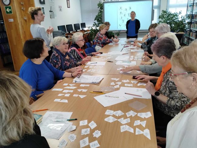 Seniorzy siedzą przy stołach, rozwiązują zadania. Prowadzące stoją przy tablicy interaktywnej.