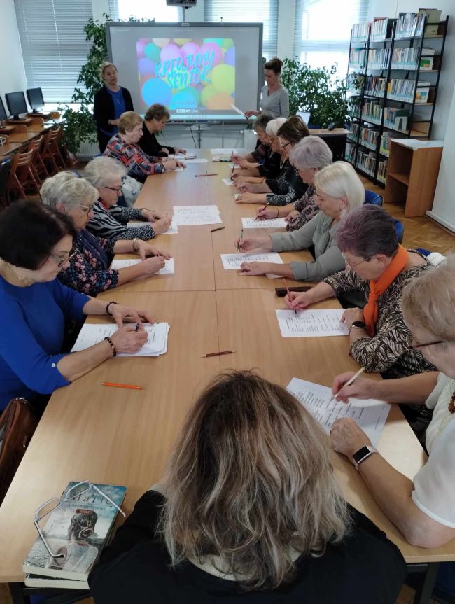 Seniorzy siedzą przy stołach, rozwiązują zadania. Prowadzące stoją przy tablicy interaktywnej.