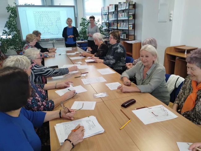 Seniorzy siedzą przy stołach, rozwiązują zadania. Prowadzące stoją przy tablicy interaktywnej.