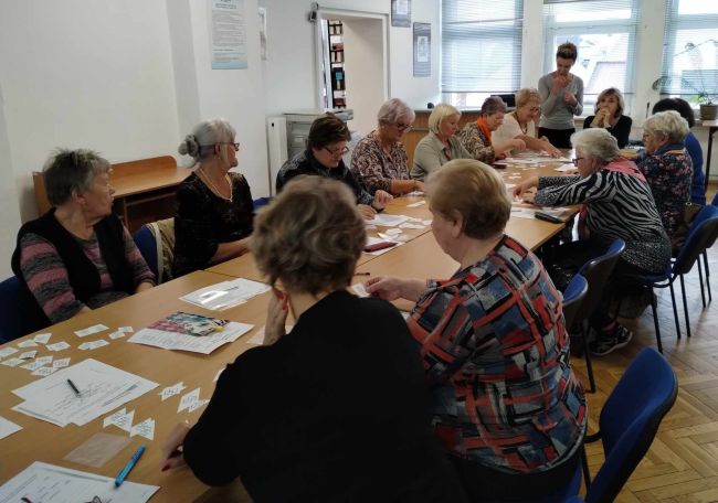 Seniorzy siedzą przy stołach, rozwiązują zadania. Prowadzące stoją przy tablicy interaktywnej.