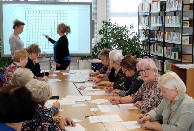 Seniorzy siedzą przy stołach, rozwiązują zadania. Prowadzące stoją przy tablicy interaktywnej.
