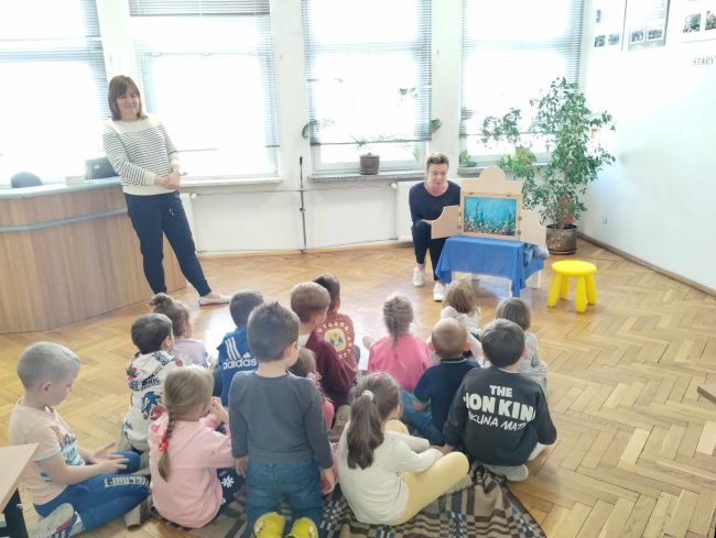 Prowadząca prezentuje bajkę  „Ogród dobrych słów” metoda Kamishibai. Dzieci siedzą na podłodze i słuchają.