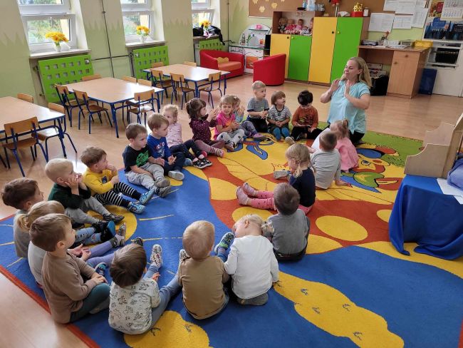 Dzieci siedzą na dywanie. Słuchają i naśladują prowadzącą zajęcia