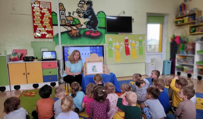 Dzieci siedzą na dywanie. Słuchają opowiadania o Myszce Pipi, prezentowanej za pomocą teatrzyku Kamishibai