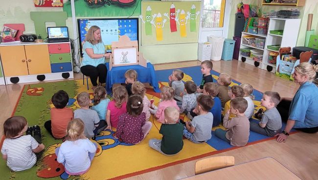 Dzieci siedzą na dywanie. Słuchają opowiadania o Myszce Pipi, prezentowanej za pomocą teatrzyku Kamishibai