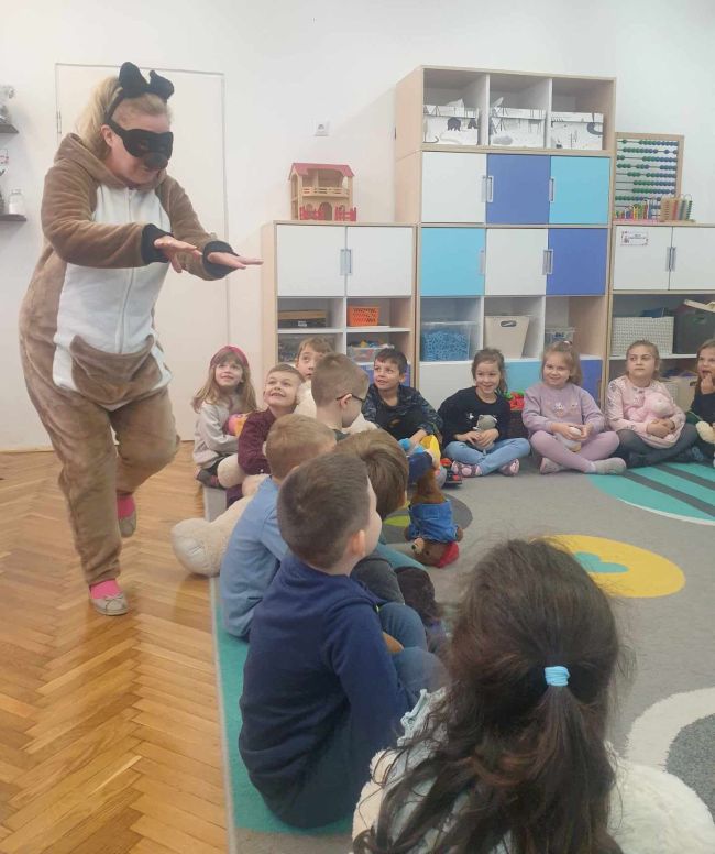 Miś Pluszowy wchodzi do sali. Dzieci siedzą na dywanie.
