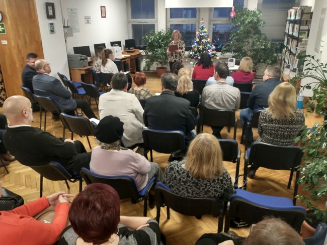 Prowadząca spotkanie. Uczestnicy wieczorku poetyckiego siedzą w sali biblioteki.