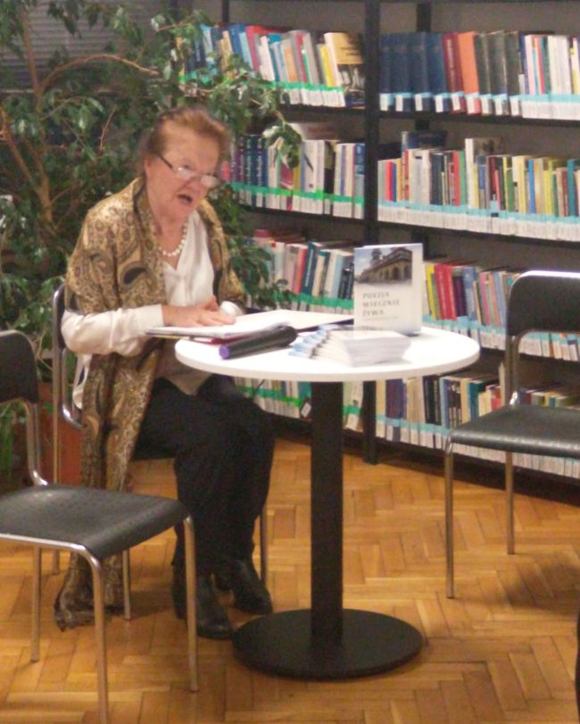 Poetka  Stanisława Różaniecka siedzi przy stoliku i odczytuje swoje utwory.