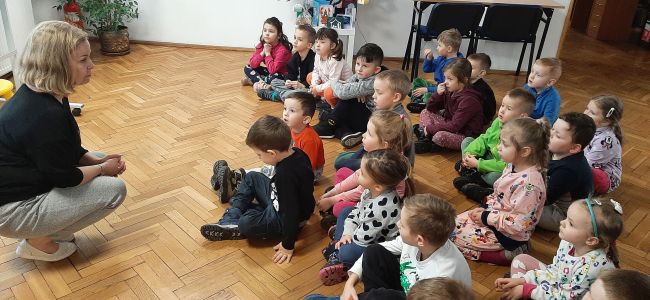 Dzieci siedzą na podłodze. Nauczyciel bibliotekarz opowiada o bibliotece.