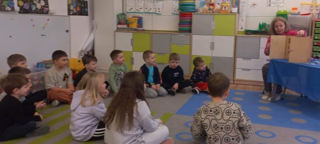 Dzieci siedzą na dywanie w sali przedszkolnej i słuchają opowieści.