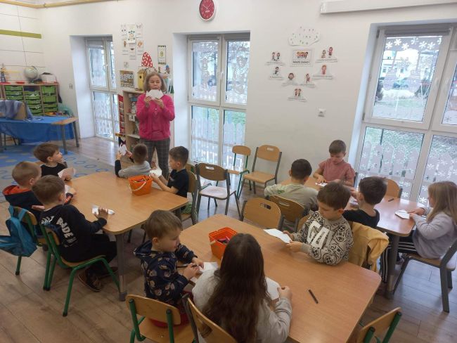 Prowadząca demonstruje, jak wykonać pieska metodą origami. Dzieci siedzą przy stolikach