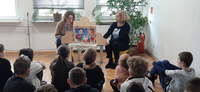 Prowadzące opowiadają bajkę”Idzie wiosna, Wielkanoc tuż, tuż…” za pomocą teatrzyku Kamishibai. Uczniowie siedzą na podłodze i słuchają
