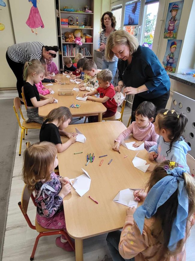 Przedszkolaki siedzą przy stolikach i z pomocą nauczycieli wykonują prace metodą origami
