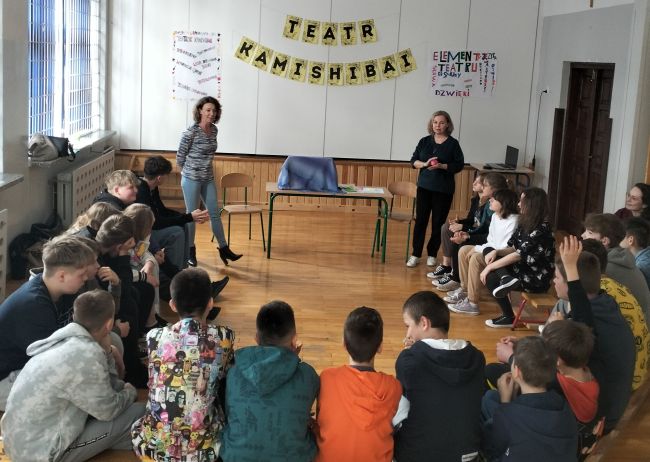 Uczniowie siedzą w półokręgu na ławeczkach gimnastycznych. Obok prowadzący zajęcia nauczyciele  bibliotekarze. Z przodu stolik ze stojącą na nim skrzynką Kamishibai. Na ścianie napis teatr Kamishibai