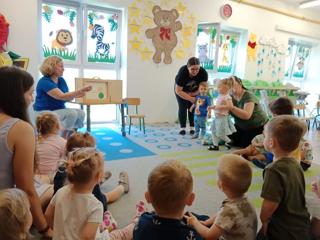 Na zdjęciu dzieci przedszkolne siedzą na dywanie oraz prowadzące prezentujące teatrzyk Kamishibai. 