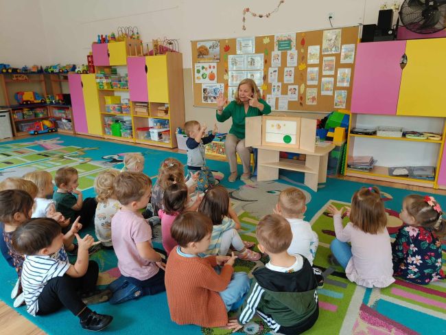 Dzieci siedzą na dywanie, oglądają i słuchają historii o Groszku, teatru Kamishibai.