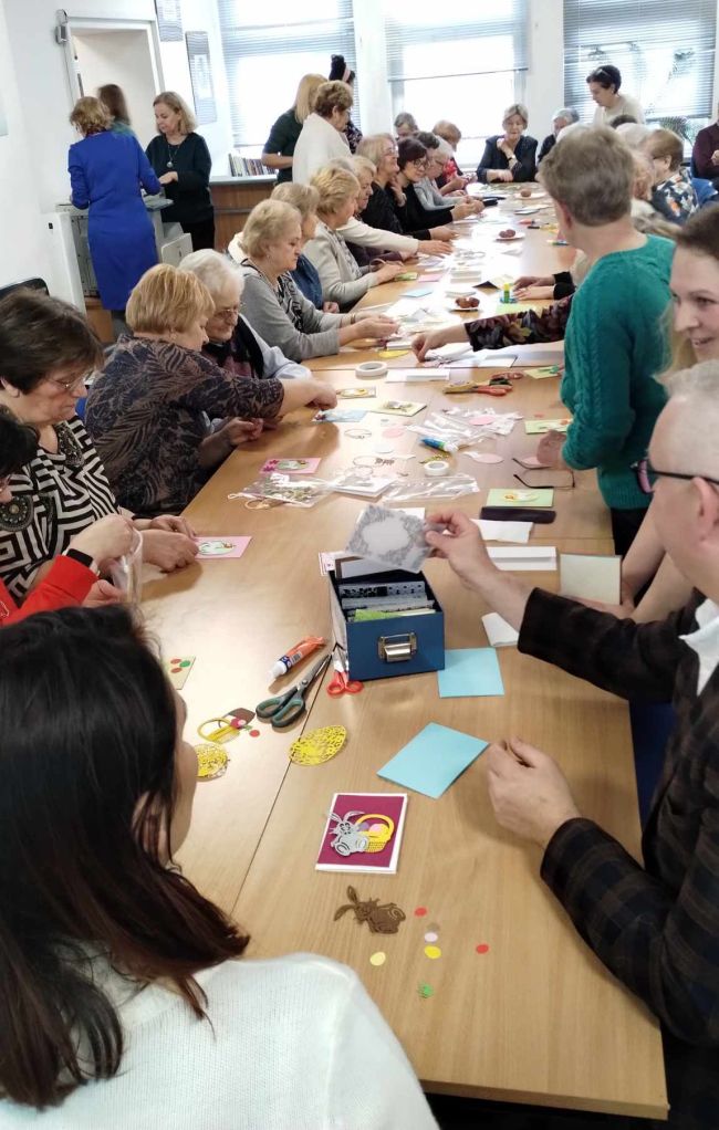 Zdjęcie zbiorowe wszystkich uczestników warsztatów. Słuchacze UTW siedzą przy stolikach i wykonują kartki metodą scrapbookingu.