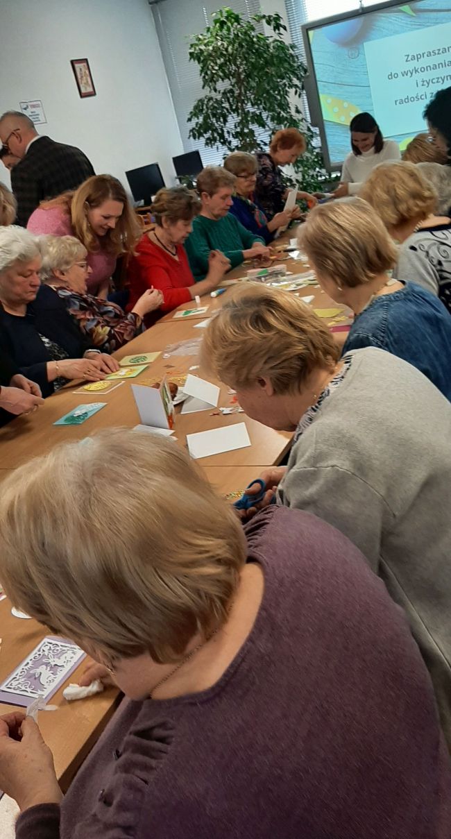 Zdjęcie zbiorowe wszystkich uczestników warsztatów. Słuchacze UTW siedzą przy stolikach i wykonują kartki metodą scrapbookingu.
