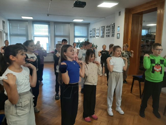 Uczniowie  stoją w czytelni i uczestniczą w zabawie prowadzonej przez nauczyciela.