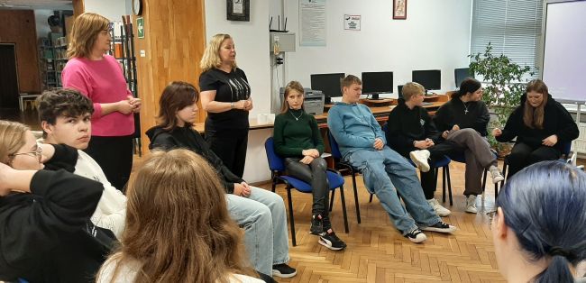 Młodzież siedzi w kole na krzesłach, nauczyciela stoją i prowadzą zajęcia.