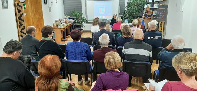 Prowadząca wita uczestników spotkania. Autorka Monika Tadra soedzi przy stoliku w centralnym miejscu.