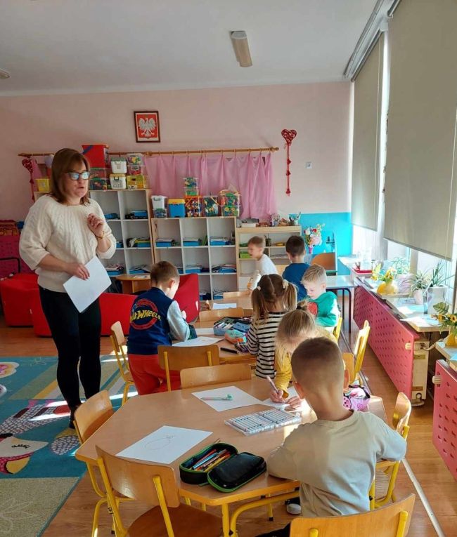 Nauczyciel wyjaśnia ćwiczenie, dzieci siedzą przy stołach i rysują.