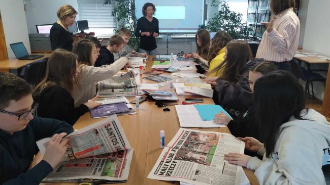 Czytelnia biblioteki. Uczniowie podzieleni na grupy siedzą przy stolikach, redagują ogłoszenie, wykorzystując w tym celu wycinki z gazet.