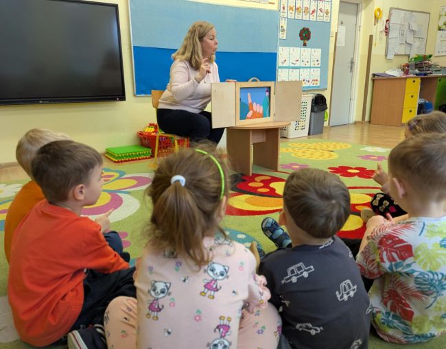 Dzieci i prowadząca siedzą na dywanie, bawią się paluszkami do piosenki