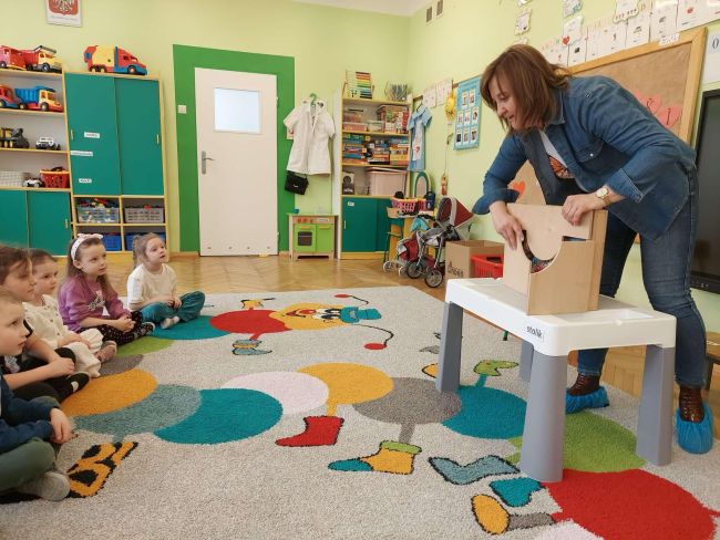 Nauczyciel prowadzi zajęcia, dzieci siedzą na dywanie i słuchają.