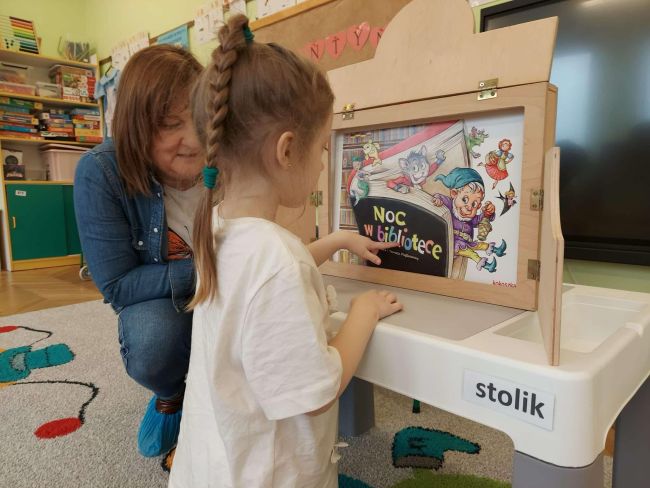 Nauczyciel prowadzi zajęcia, dziewczynka stoi przy skrzynce kamishibai i czyta.