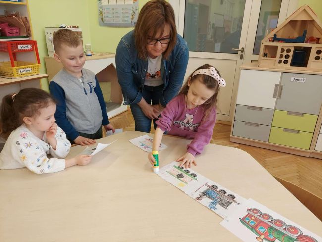 Dzieci stoją przy stoliku  przy i przyklejają swoje wagoniki.