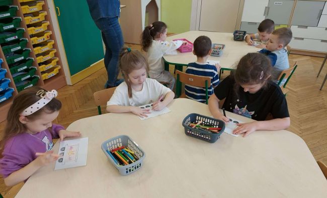 Przedszkolaki siedzą przy stołach i malują prace plastyczne.